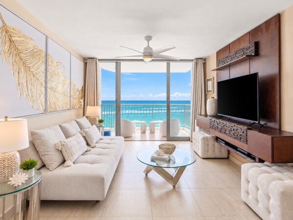 A modern living room with a sofa, coffee table, and TV, overlooking a balcony with a view of the ocean.