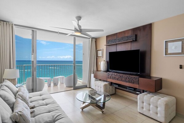A modern living room with a large TV, comfortable seating, and a balcony with a view of the ocean.