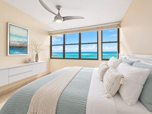 A bedroom with a large bed, a ceiling fan, a window with a view of the ocean, and a white dresser.