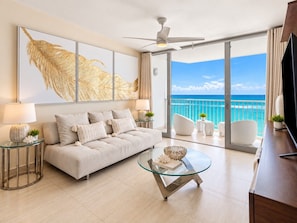 A modern living room with a sofa, coffee table, and chairs, a large window with a view of the ocean, and a ceiling fan.