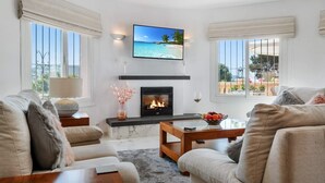 A living room with a fireplace, a television mounted above it, and a view of the outdoors through large windows.