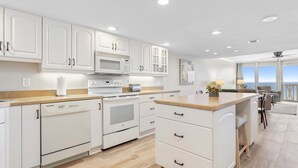 Een moderne keuken met witte kasten, een magnetron, een oven en een vaatwasser. Er is een houten aanrecht en een klein eiland met zitplaatsen. De keuken grenst aan een balkon met uitzicht op de zee.