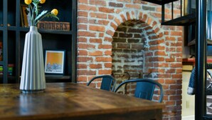 A wooden table with a white vase containing yellow flowers, a brick wall with an arched window, and metal chairs.