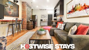 A modern living room with a sofa, coffee table, and chairs. There is a brick wall, a fireplace, and a kitchen area in the background.