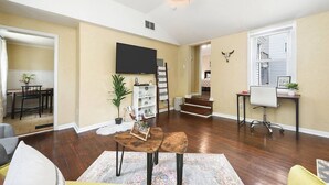 A spacious living room with hardwood flooring, a flat-screen TV mounted on the wall, a staircase leading to another level, and a desk with a chair. - 1210 James St. #2 · Free Off Street Parking! (Pittsburgh)