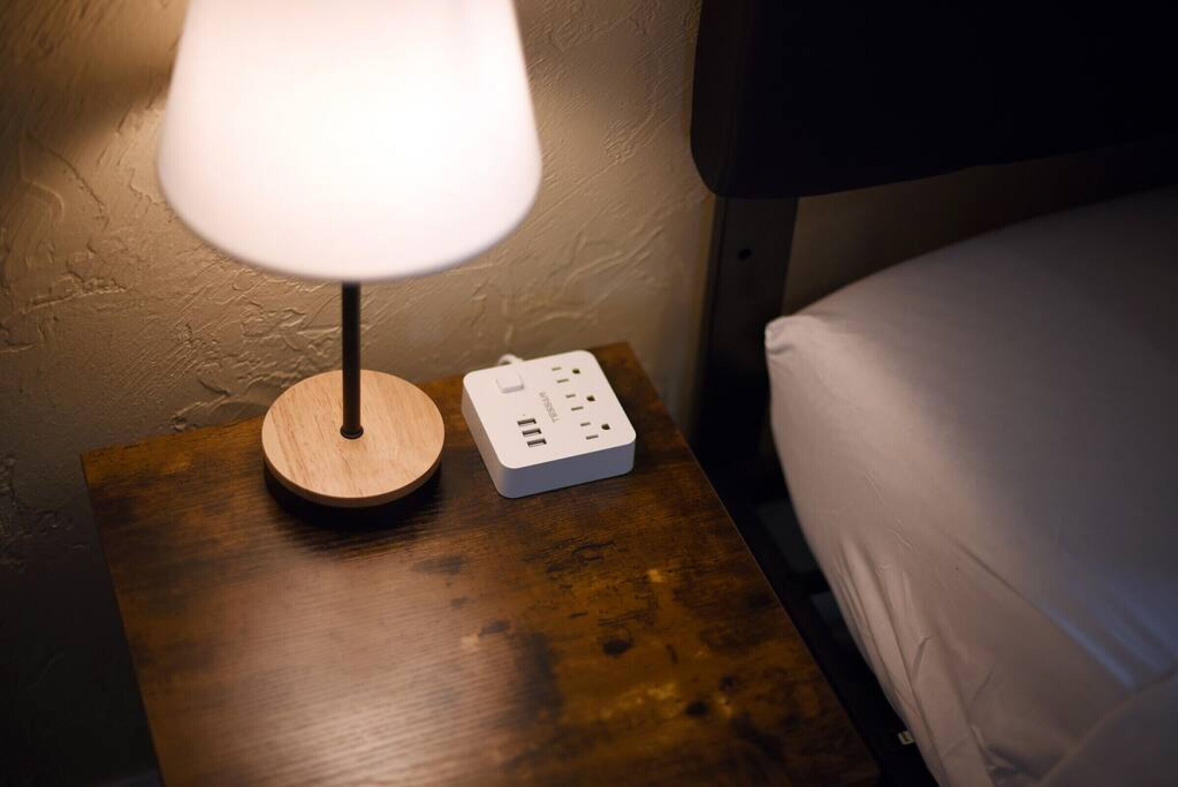 A wooden bedside table with a white lamp and a power strip.