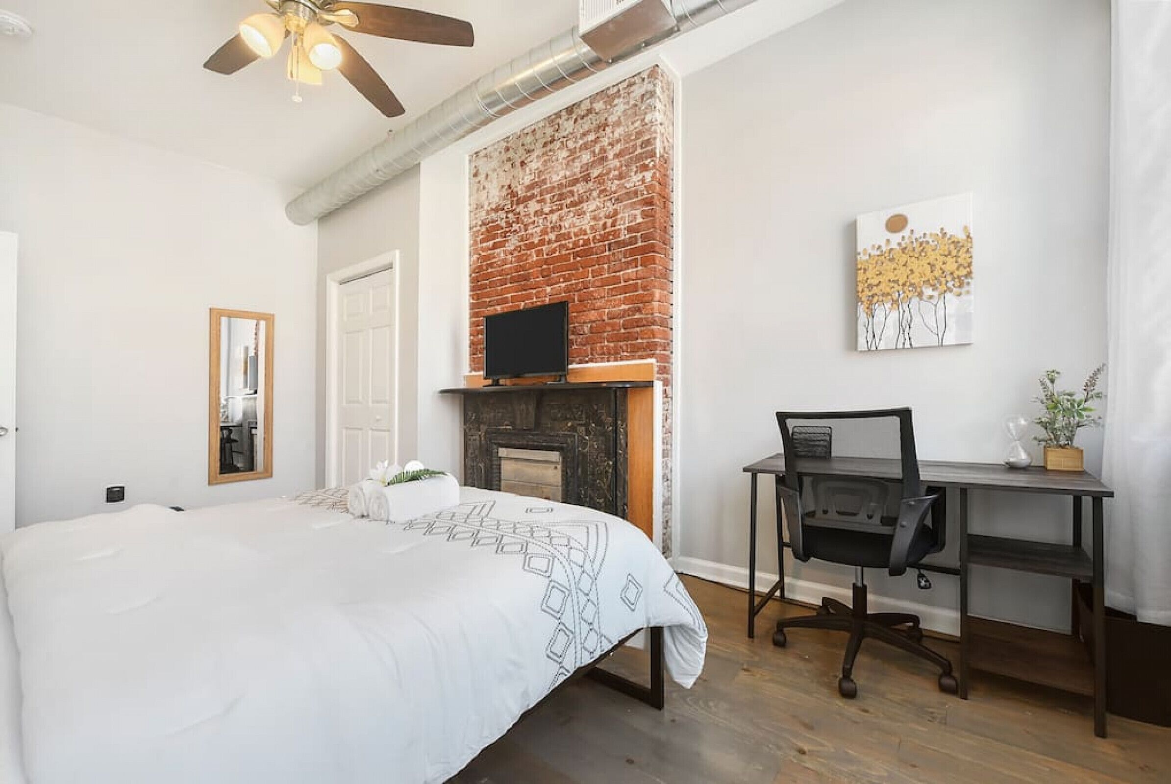 A bedroom with a brick fireplace, a bed with a white comforter, a desk with a computer, and a ceiling fan.