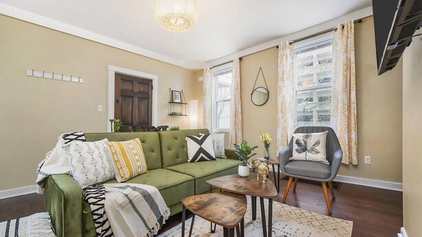 Ein Wohnzimmer mit einem grünen Sofa, einem Couchtisch und einem Sessel. Außerdem ist ein Kleiderständer und ein Fernseher an der Wand zu sehen.