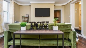 Ein Wohnzimmer mit einem grünen Sofa, einem Tisch mit der Aufschrift 'PGH', einem Flachbildfernseher und einer Topfpflanze.