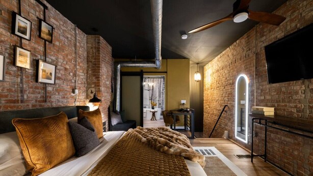 Une chambre moderne avec des murs en briques apparentes, un grand lit recouvert d’une couverture à motifs, un ventilateur de plafond et un téléviseur à écran plat fixé au mur.