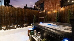 Un bain à remous dans un jardin, avec une clôture en bois, des guirlandes lumineuses et de la neige au sol.