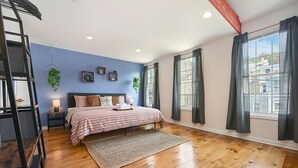 A bedroom with a bed, a nightstand, a lamp, a hanging plant, and a view of the outside through large windows. - 1207 Ocala - Boat House - North Side (Pittsburgh)