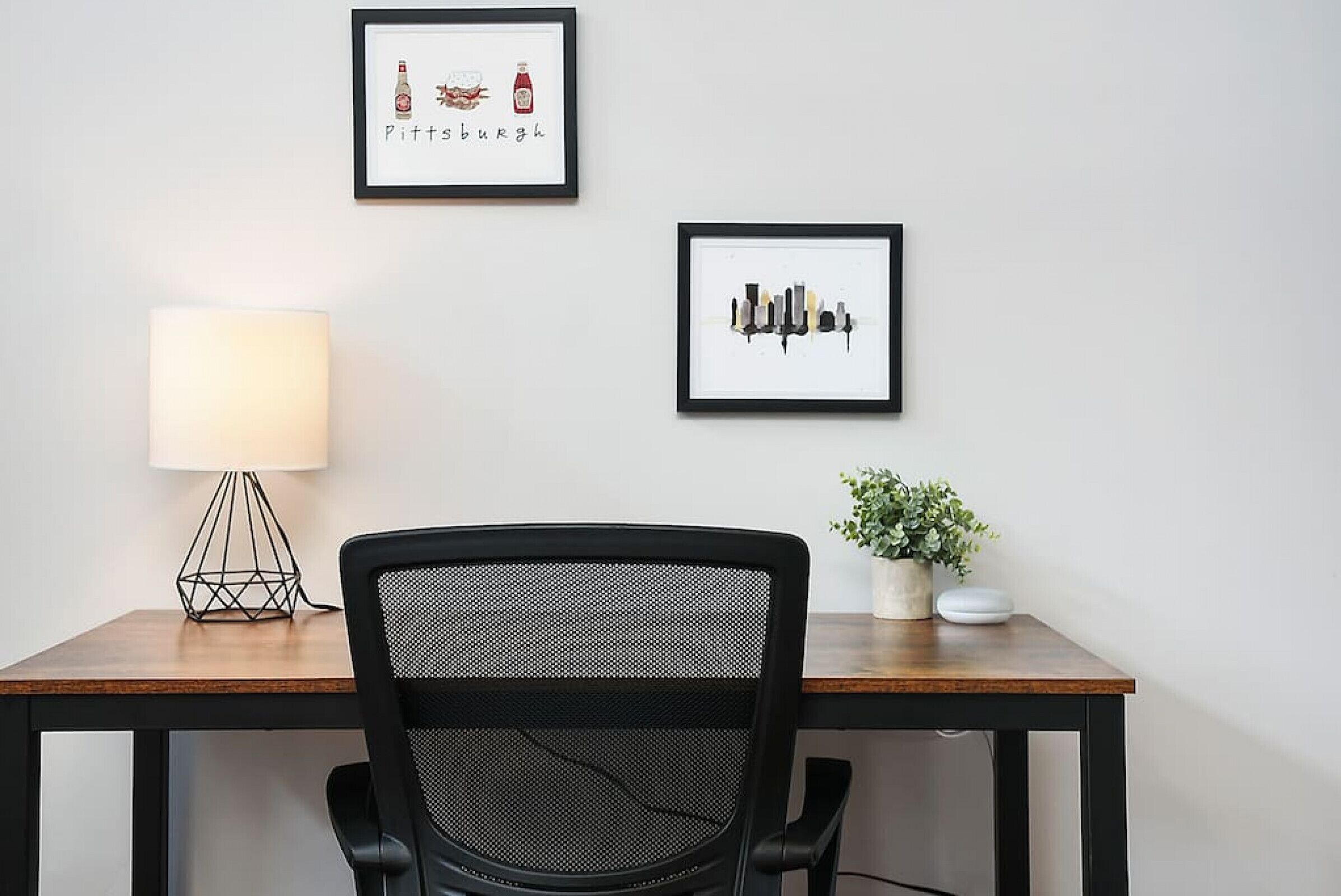 A workspace with a desk, chair, lamp, framed artwork, and a potted plant.