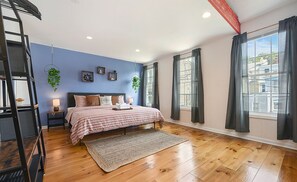 A bedroom with a bed, a nightstand, a lamp, a hanging plant, and a view of the outside through large windows. - 1207 Ocala - Boat House - North Side (Pittsburgh)