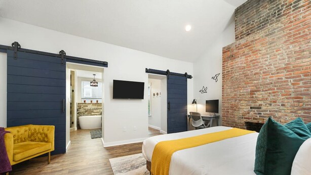 A modern hotel room with a large bed, a yellow armchair, a desk, and a brick wall. - 190.5 44th St. - Lawrenceville (Pittsburgh)