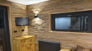 A compact room with a wooden wall, a mounted TV, a window with blinds, a radiator, and a wooden cabinet. - Studio Cabin 25m² at the foot of the slopes (Val Thorens)