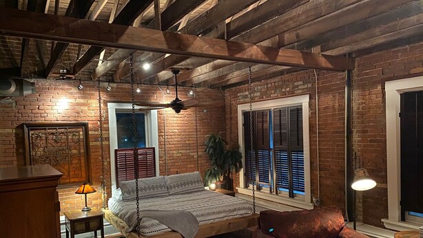 A cozy room with exposed wooden beams, a bed with a swing, a red sofa, and a brick wall.