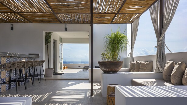 Un área de estar exterior moderna con vista al mar, que incluye un bar, sillones de descanso y una planta grande en maceta.