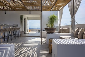Un área de estar exterior moderna con vista al mar, que incluye un bar, sillones de descanso y una planta grande en maceta.