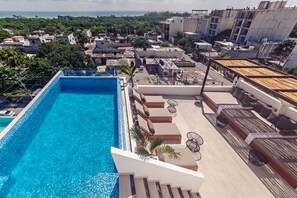 A rooftop pool with clear blue water, surrounded by lounge areas with seating and umbrellas, overlooking a densely built urban area with visible greenery and buildings.