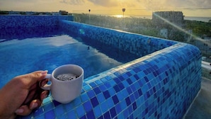 Una persona sostiene una taza cerca de una piscina infinita con azulejos azules y vista al atardecer.