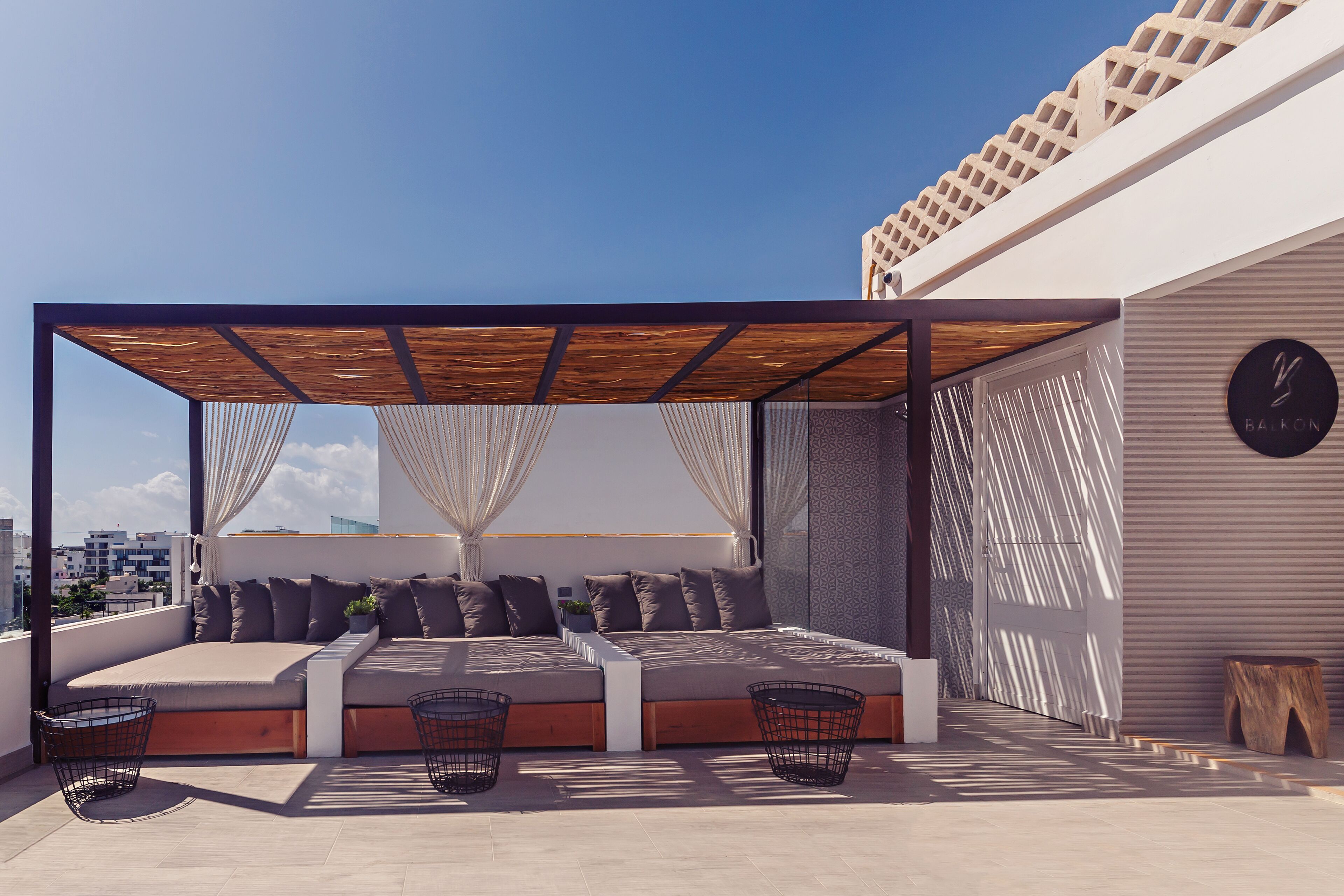 A rooftop terrace with a wooden pergola, cushioned seating, and a view of the cityscape.