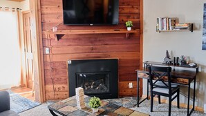 A living room with a fireplace, a television mounted on a wooden wall, a bookshelf, and a dining area with a table and chairs. - Cozy Condo at Summit- 103D (Snowshoe)