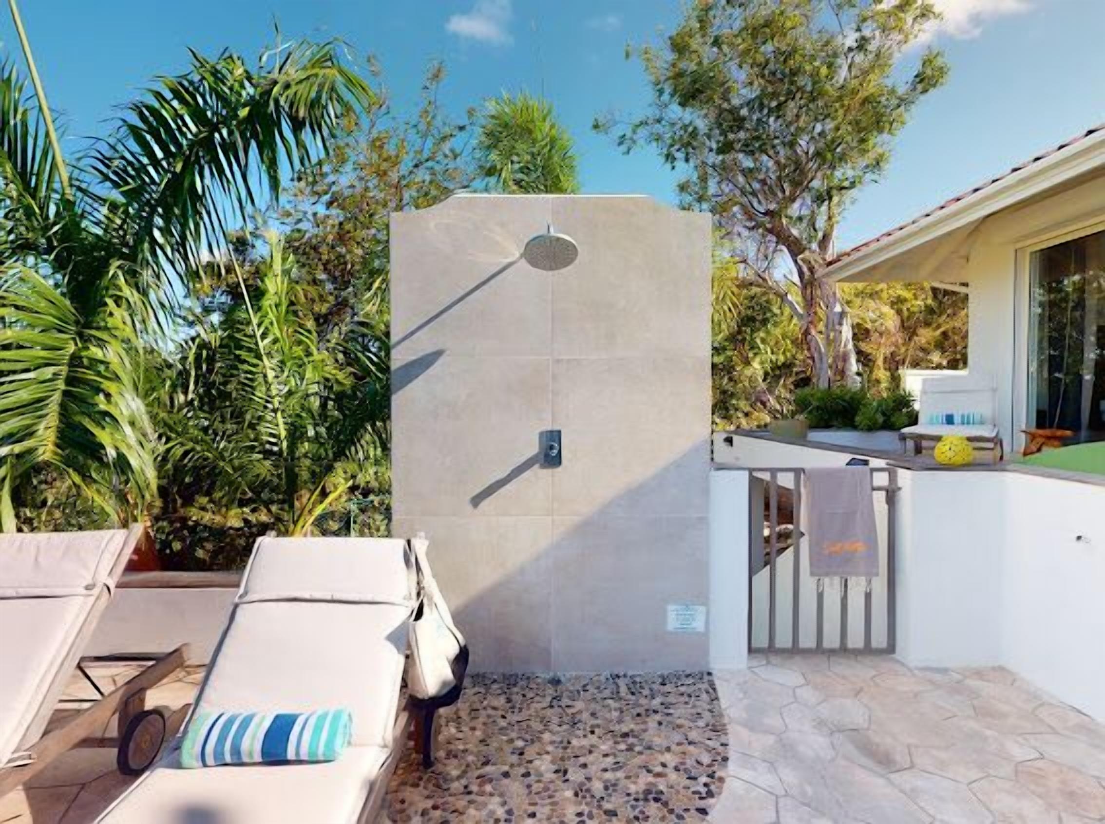 Un espace de douche extérieur moderne, doté d’un sol en pierre, entouré d’une végétation luxuriante et agrémenté d’un coin salon au bord de la piscine.