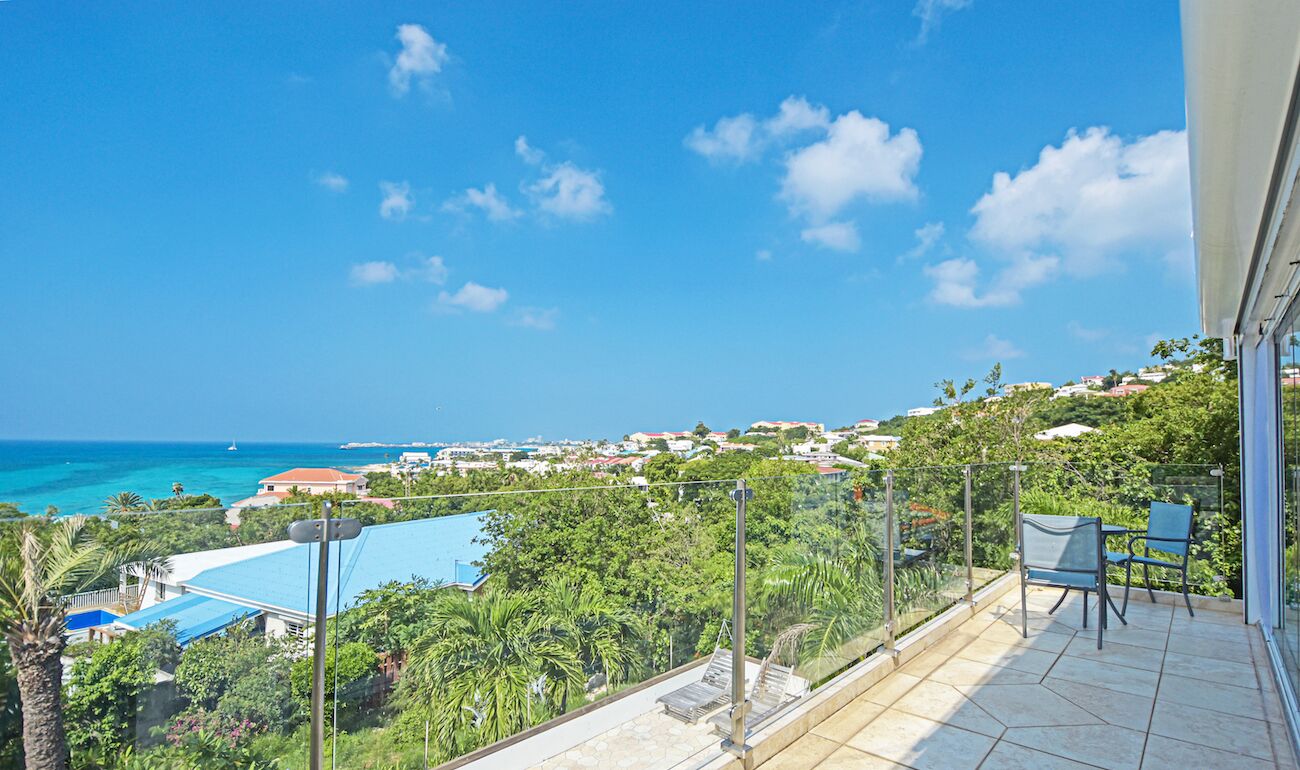 A balcony with a view of the sea and a swimming pool, surrounded by greenery.