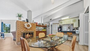 A spacious living room with a glass-top dining table surrounded by wicker chairs, a brick fireplace, and a view of the kitchen. - Waterfront! Firepit~3 King bedrms~playroom~kayaks~near restaurants (Hot Springs)