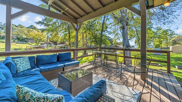 A covered outdoor seating area with a sofa and chairs, a table, and a view of a grassy yard and trees. - Waterfront! Firepit~3 King bedrms~playroom~kayaks~near restaurants (Hot Springs)