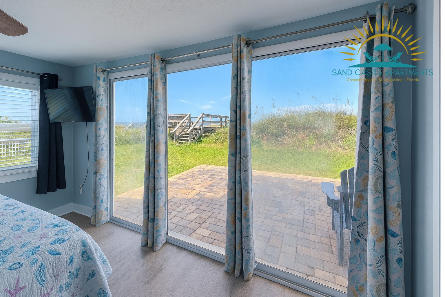 A room with a large sliding glass door leading to an outdoor patio with a wooden deck and a view of a beach and a wooden staircase.