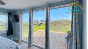 Een kamer met een grote schuifglazen deur die uitkomt op een terras met een houten terras en uitzicht op een strand en een houten trap.