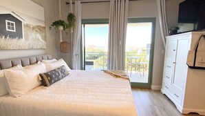 A bedroom with a bed, a ceiling fan, a large window with curtains, a view of the outdoors, and a white dresser.