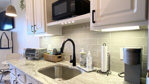 A modern kitchen with white cabinets, a stainless steel sink, a microwave, a coffee maker, and a hanging light fixture.