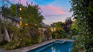 Un área de piscina con una pequeña terraza, rodeada de abundante follaje e iluminación con luces colgantes.
