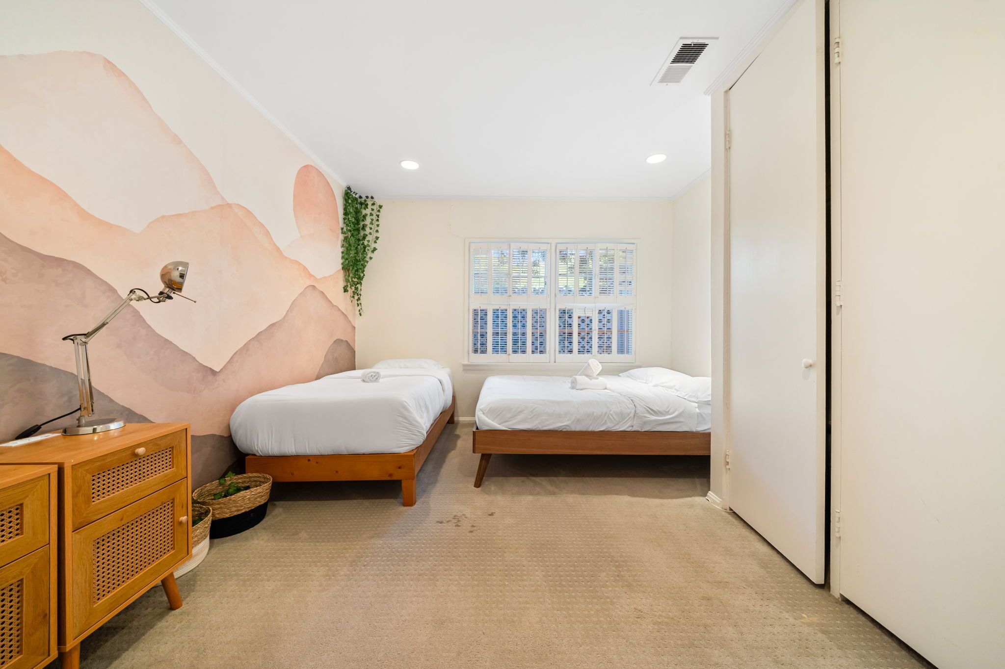 Un dormitorio con dos camas, una mesita de noche de madera, una lámpara de pared y una ventana con persianas.