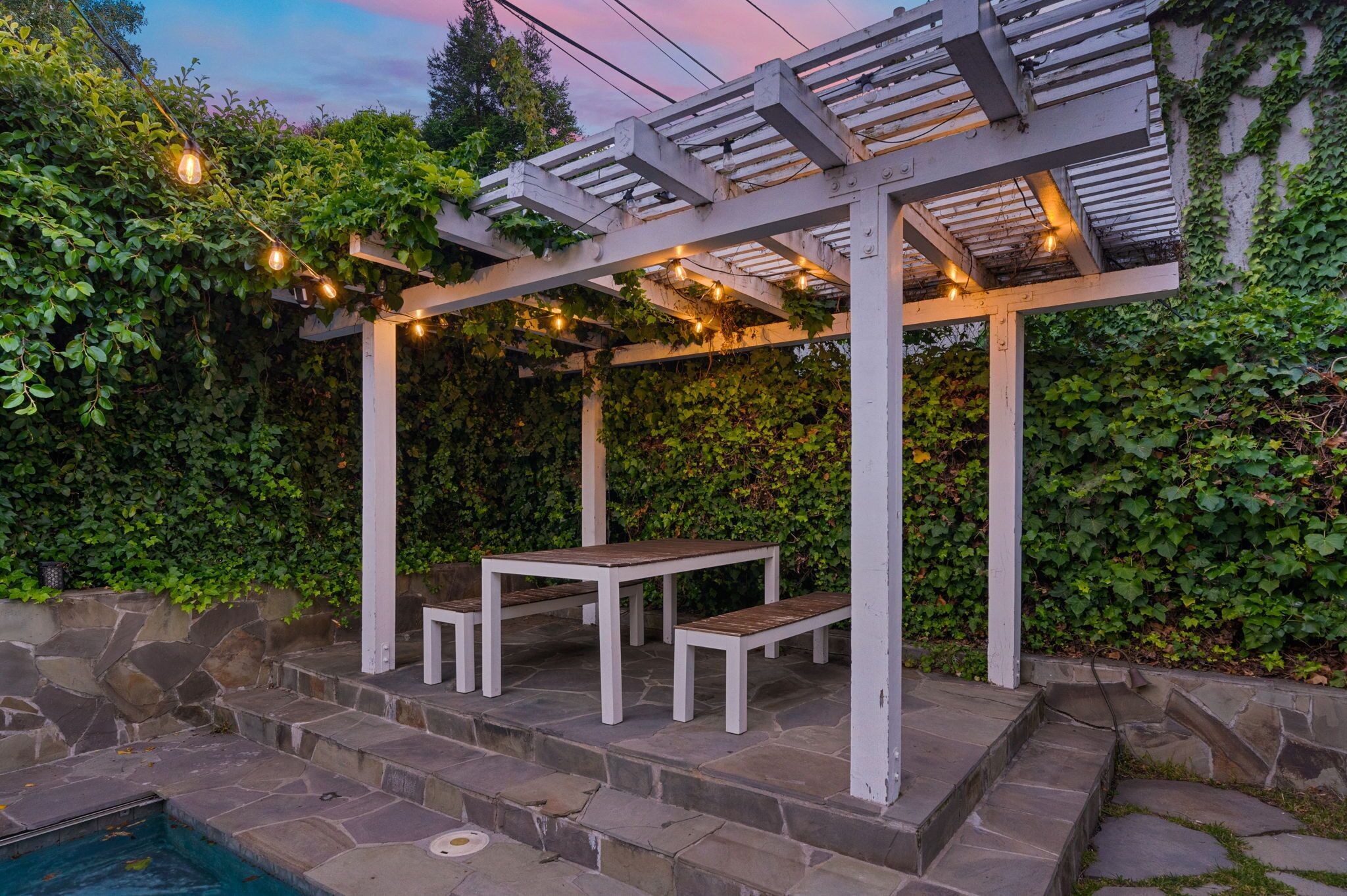 A covered outdoor seating area with stone steps leading to a pool, surrounded by lush greenery.