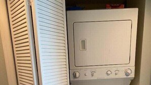 Ein kleiner, weißer Hauswirtschaftsraum mit integrierter Waschmaschine und Trockner, einem kleinen Fenster und einer Tür mit Lamellen.