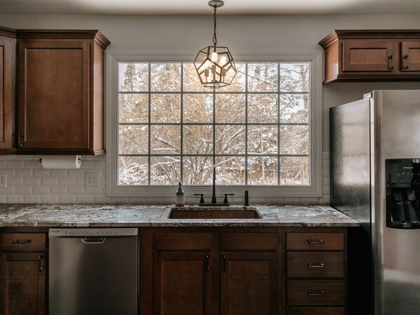 A kitchen with a stainless steel refrigerator, wooden cabinets, a window with a view of snow-covered trees, and a pendant light. - The Hermantown Hangout (Hermantown)