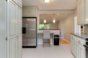 Una cocina con gabinetes blancos, un refrigerador de acero inoxidable, una silla de madera y un cubo de basura.