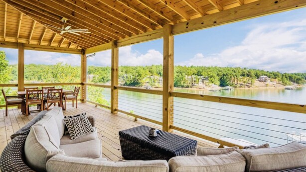 Eine überdachte Terrasse mit Holzboden, Deckenventilator, einer Couch, einem Tisch und Stühlen. Durch die Geländer blickt man auf einen See und grüne Landschaft.