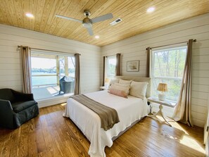 Een slaapkamer met een groot bed, houten vloeren, een plafondventilator en uitzicht op een waterpartij door een raam.