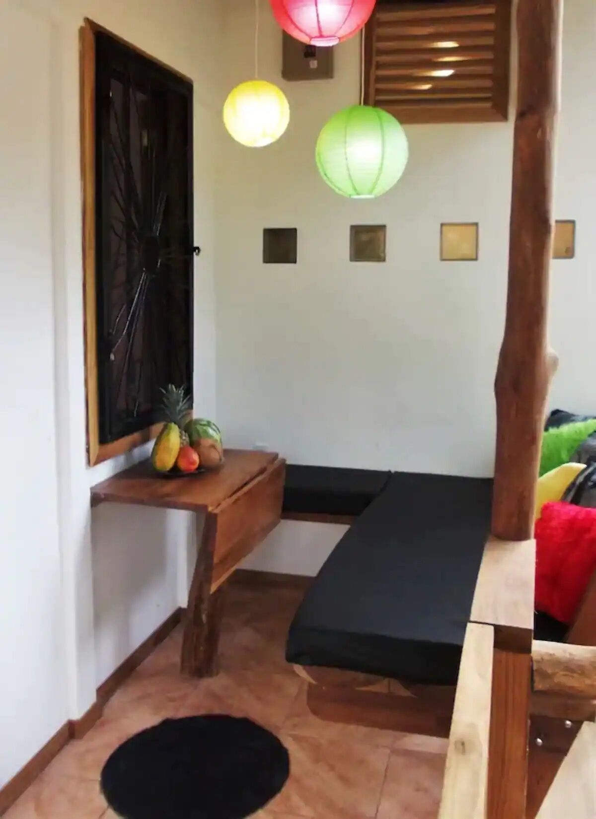 Ein kleines Zimmer mit einem Bett, einem Holztisch mit Früchten, einer schwarzen Matte und einer Wand mit drei kleinen Bilderrahmen.