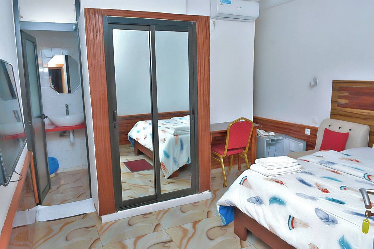 A hotel room with a bed, a desk, a chair, a mirror, and an air conditioning unit.