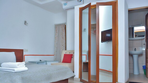A hotel room with a bed, a nightstand, a chair, a mirror, a television, and a bathroom with a sink and toilet. - Florence Hotel Firenze Yamoussoukro (Yamoussoukro)