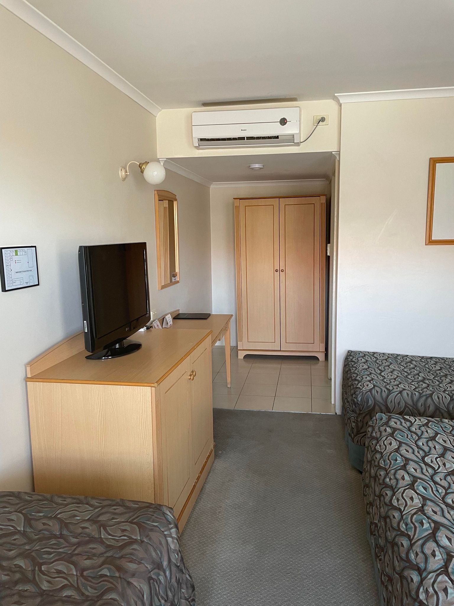 Una habitación con televisión sobre un escritorio de madera, un armario, una cama con ropa de cama estampada y un equipo de aire acondicionado en la pared.