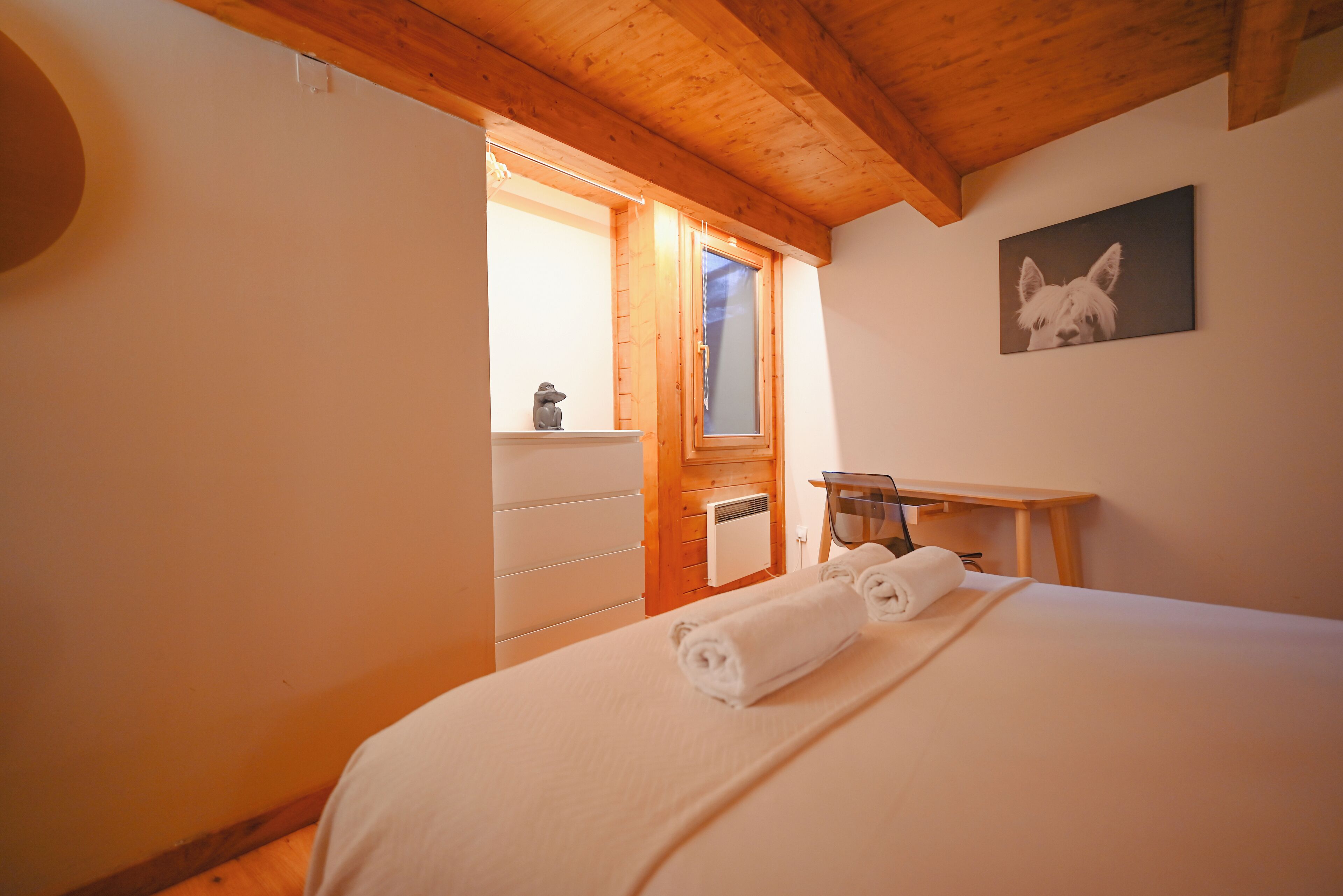 A bedroom with a wooden ceiling, a bed with white linens, a wooden desk, and a framed picture on the wall.