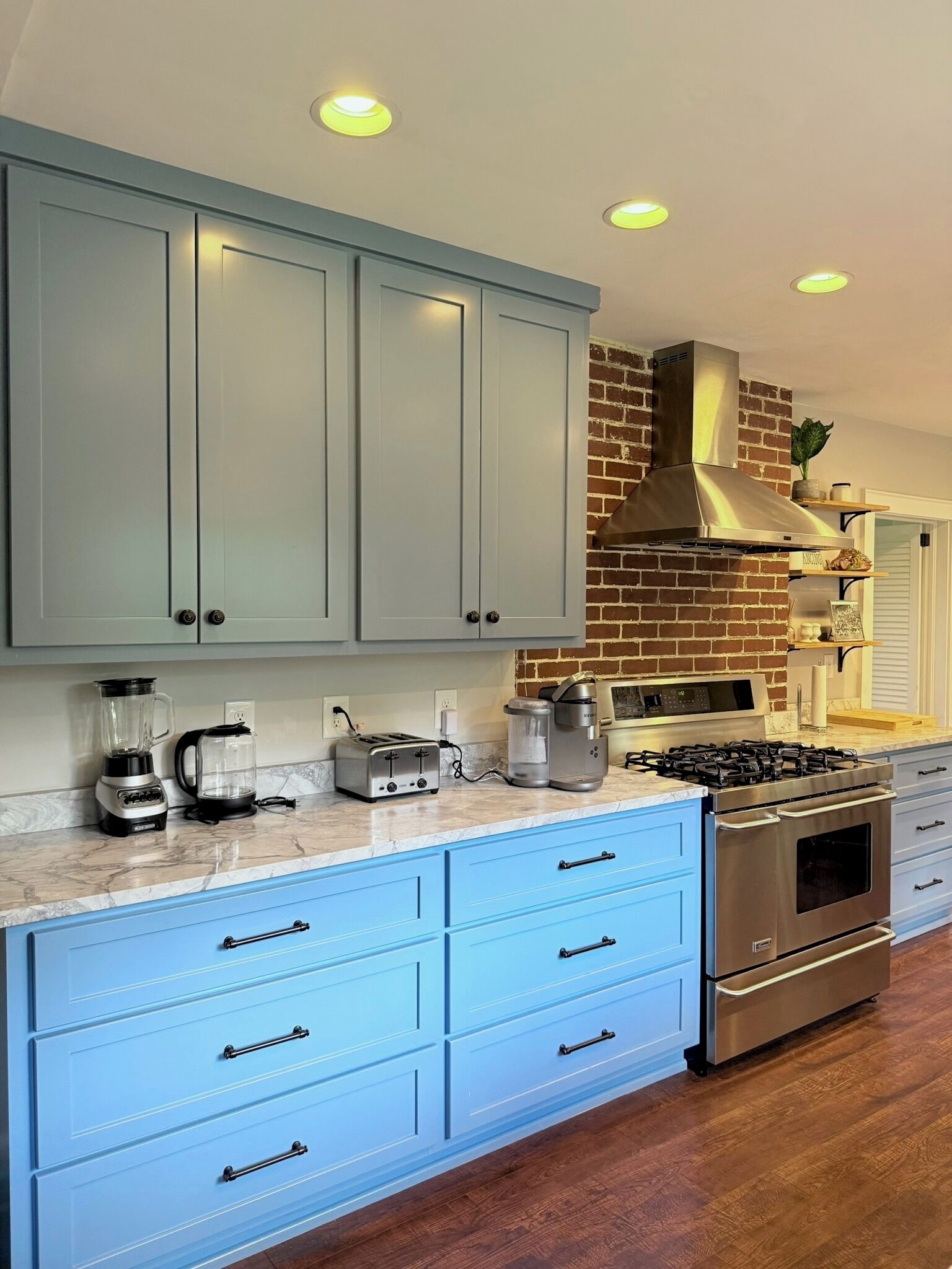 A kitchen with light blue cabinets, a brick wall, stainless steel appliances, and wooden flooring.