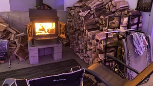 Una habitación con un estufa de leña, un montón de leña y una silla con una manta.
